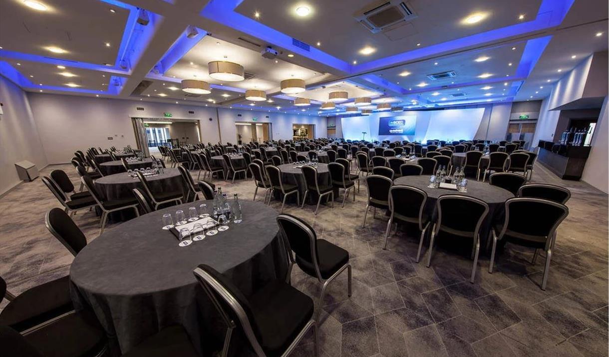 Showing the interior of the Birmingham Conference and Events Centre, the venue for the National Planning Conference. Many circular tables and chairs facing a stage with blue lighting